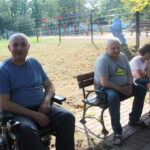 mężczyzna na wózku patrzy w aparat, obok grupa osób w parku, rozmawiają, uśmiechają się