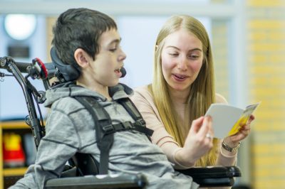 A caregiver is playing with a boy with a physical disability - he is sitting in a wheelchair and smiling happily.